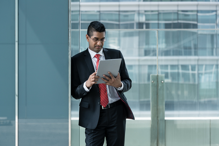 Man in a business suit leaning against a glass column reading a document.